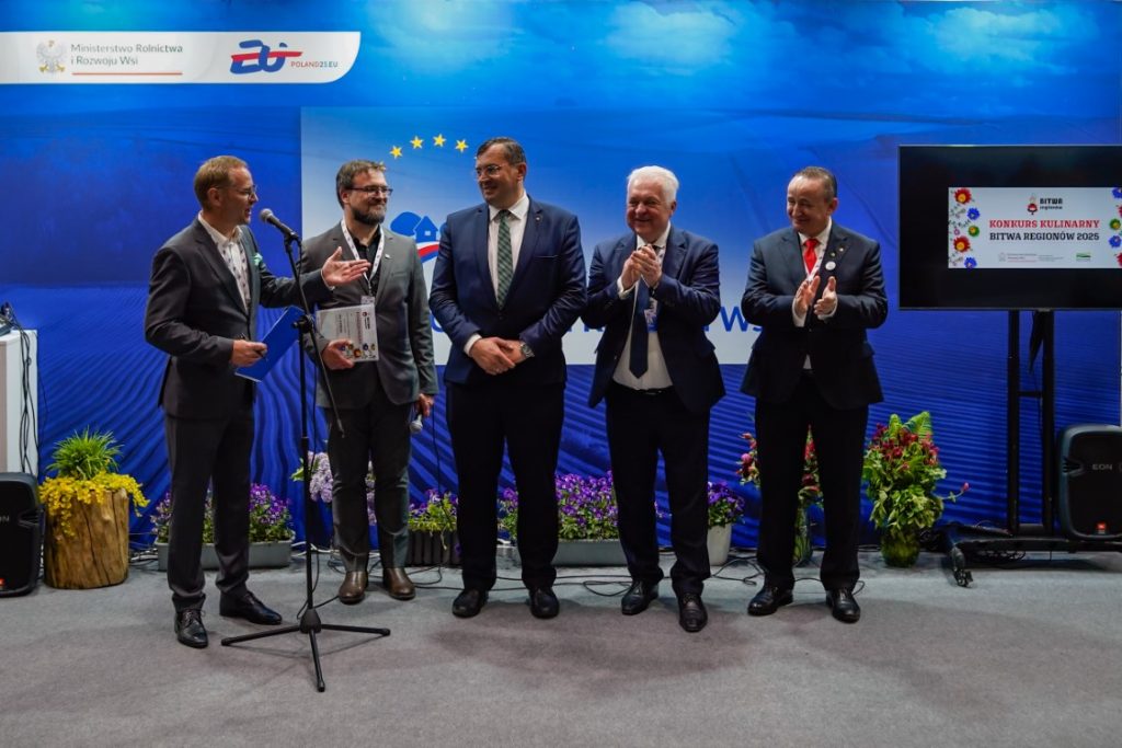 Sekretarze stanu w Ministerstwie Rolnictwa i Rozwoju Wsi – Jacek Czerniak i Stefan Krajewski – wraz z innymi uczestnikami wydarzenia pozują przy ściance promocyjnej. Pierwsza osoba z lewej przemawia do mikrofonu i wskazuje pozostałych. Zdjęcie wykonane podczas I etapu X edycji „Bitwy Regionów”.