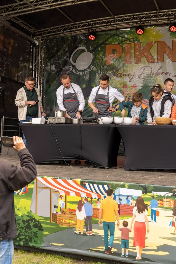 <p><span style="color: #000000;"> Na scenie dwaj kucharze gotują z dziećmi przy stołach z akcesoriami kuchennymi, po lewej stoi konferansjer z mikrofonem. </span></p>