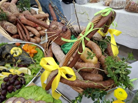 Kosz wypełniony różnymi rodzajami tradycyjnych wędlin i kiełbas, ozdobiony zielonymi gałązkami oraz żółtymi i zielonymi wstążkami. Wokół widoczne są inne potrawy wielkanocne, m.in. sałatki, owoce i dekoracje stołu.