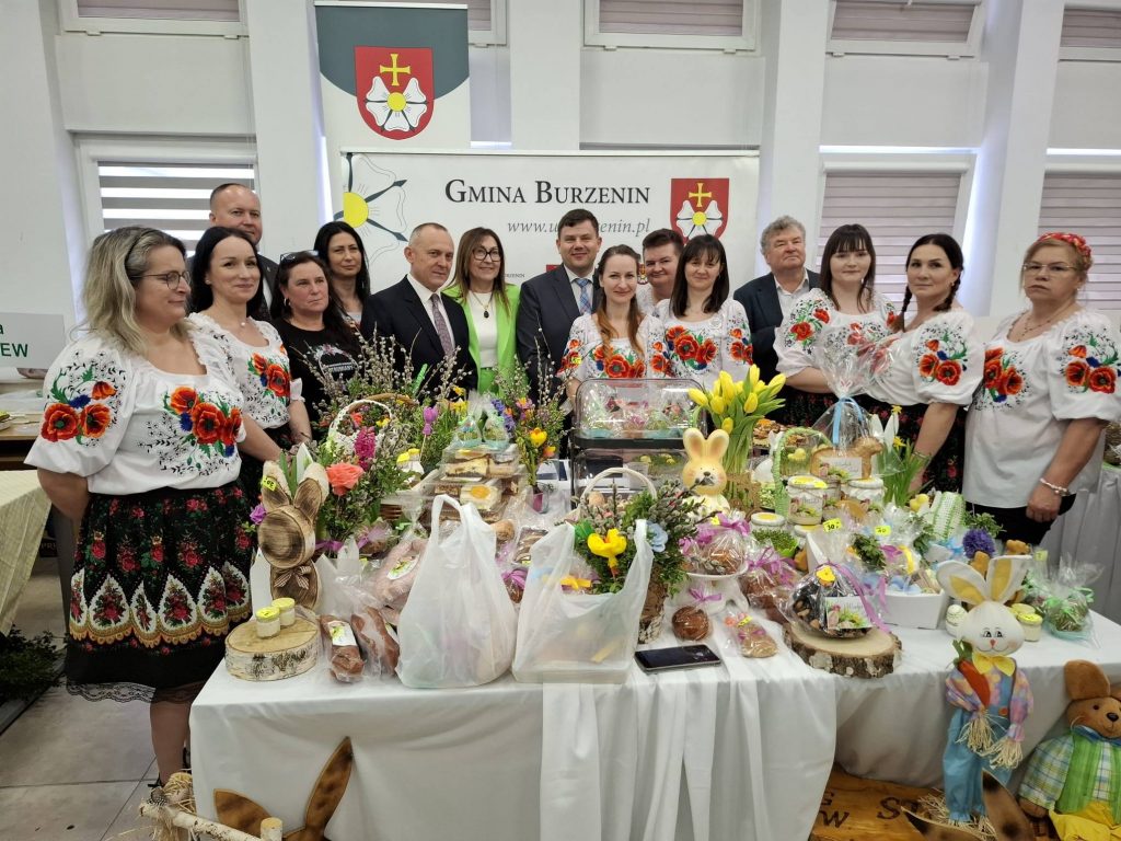 Jarosław Janiak – Burmistrz Gminy Burzenin, Iwona Mamzer – Dyrektor ŁODR w Kościerzynie, Przemysław Jaszczak – Dyrektor KOWR w Łodzi oraz przedstawicielki Koła Gospodyń Wiejskich podczas wydarzenia, stoją przy stole z tradycyjnymi potrawami wielkanocnymi i rękodziełem.