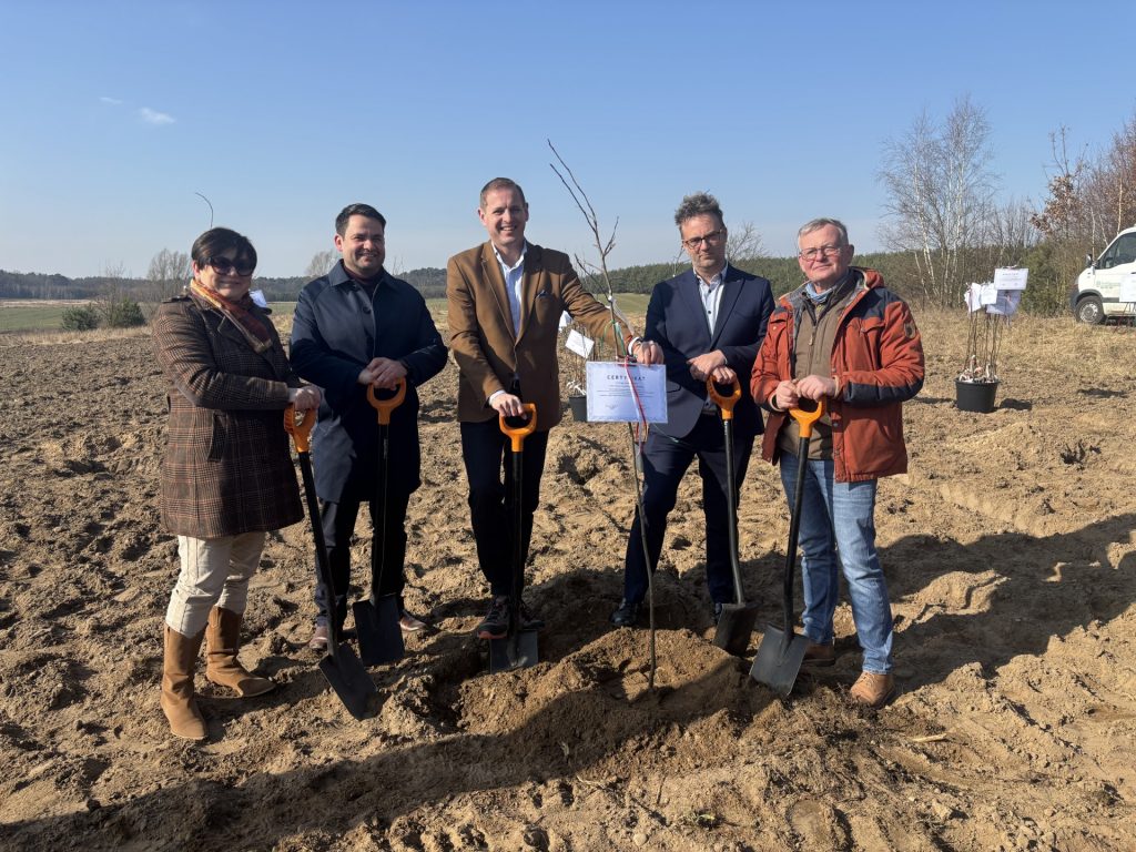 Pięć osób stoi na polu przy świeżo posadzonej jabłoni odmiany reneta landsberska i trzyma łopaty po zakończeniu symbolicznego sadzenia drzewa.