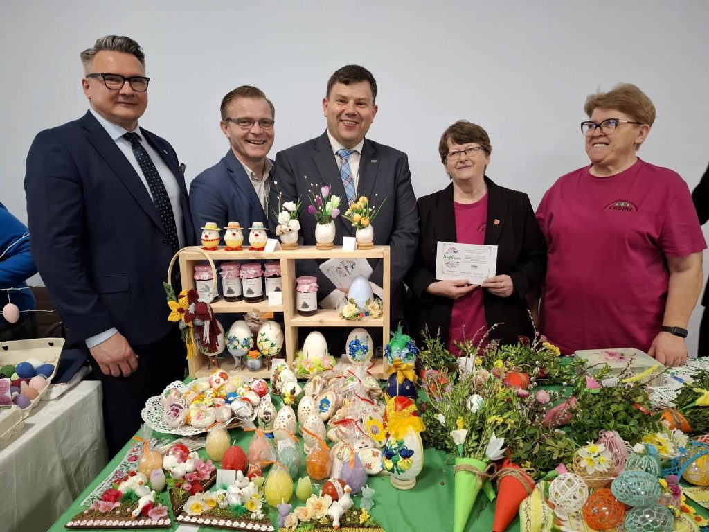 Przedstawiciele KOWR oraz uczestnicy wydarzenia stoją przy stole z tradycyjnymi wyrobami wielkanocnymi podczas Wystawy Tradycyjnych Sieradzkich Stołów Wielkanocnych; na stole znajdują się ręcznie zdobione pisanki, dekoracje świąteczne oraz lokalne produkty, promowane w ramach inicjatywy #KupujŚwiadomie.