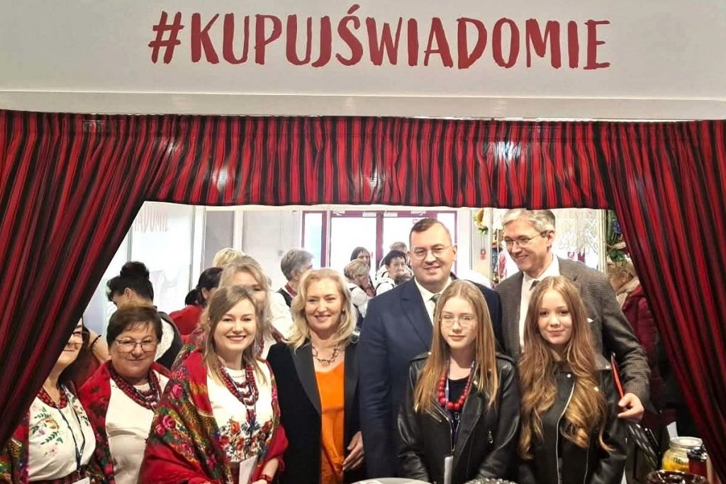 Uczestnicy Targi Agro&Active Travel 2026 stoją przy stoisku z regionalnymi potrawami pod hasłem „#KUPUJŚWIADOMIE”. Wśród nich są Małgorzata Wilk-Grzywna, Stefan Krajewski oraz Adam Jarubas, w otoczeniu przedstawicielek Kół Gospodyń Wiejskich i młodych uczestniczek wydarzenia. Na pierwszym planie widoczne są tradycyjne produkty i potrawy regionalne.
