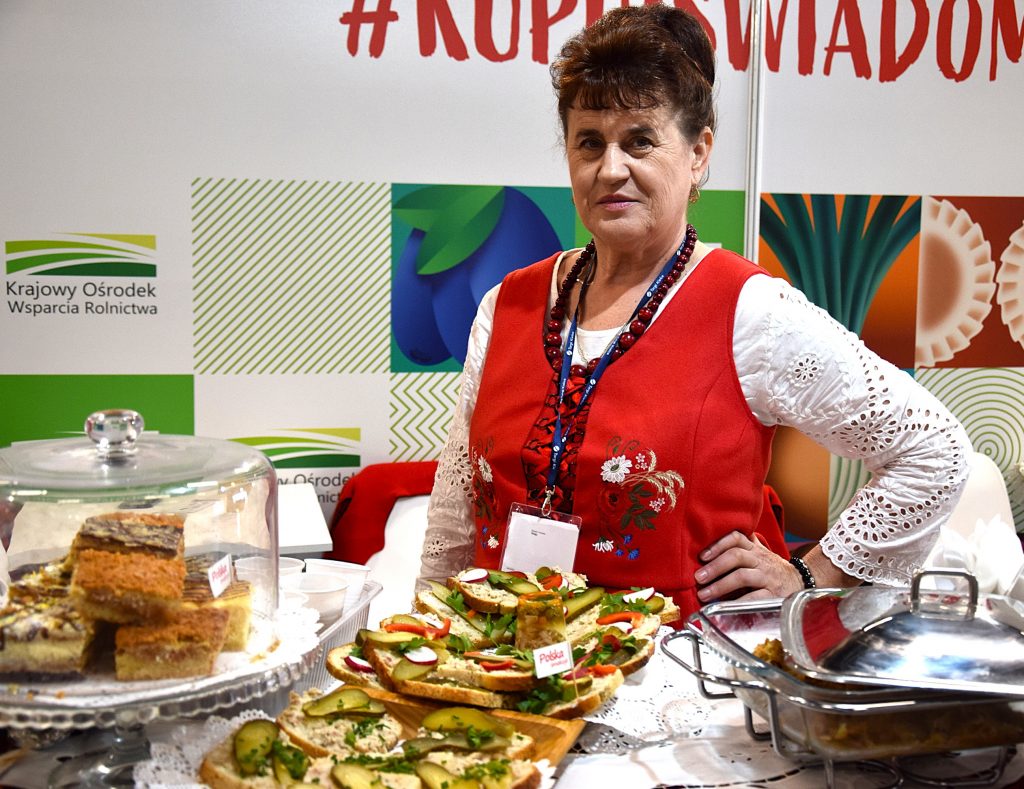 Przedstawicielka Koła Gospodyń Wiejskich w czerwonym stroju ludowym prezentuje kanapki i przekąski na stole podczas Targi Agro&Active Travel 2026.