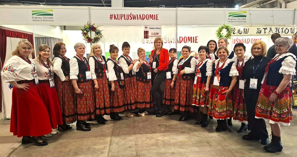 Justyna Kasprzyk stoi przy stoisku z regionalnymi potrawami i pozuje z przedstawicielkami Koła Gospodyń Wiejskich podczas wydarzenia Targi Agro&Active Travel 2026.